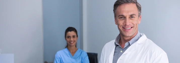 A doctor is smiling while a nurse or assistant also smiles in the background